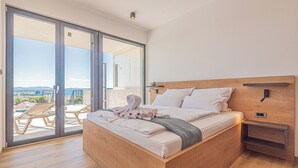 A modern bedroom with a wooden bed, a large sliding glass door leading to a balcony, and a small bedside table. - Aliara Panorama 1 
Luxusvilla (Vodice)