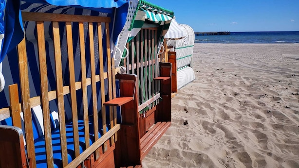 En trækonstruktion ved stranden med en blå markise, en stribet parasol og hav og mole i baggrunden.