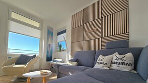 A modern living room with a grey sofa, a small round table, and a chair. There is a decorative wall panel with a logo and a painting of a sailboat.