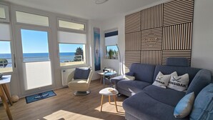 A modern living room with a sofa, a chair, a small table, and a view of the sea through large windows.