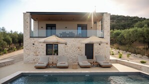 A modern house with a stone exterior, a pool, and two lounge chairs with umbrellas.