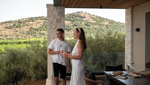 A couple standing on a balcony overlooking a vineyard, with a dining area and a stone pillar visible.