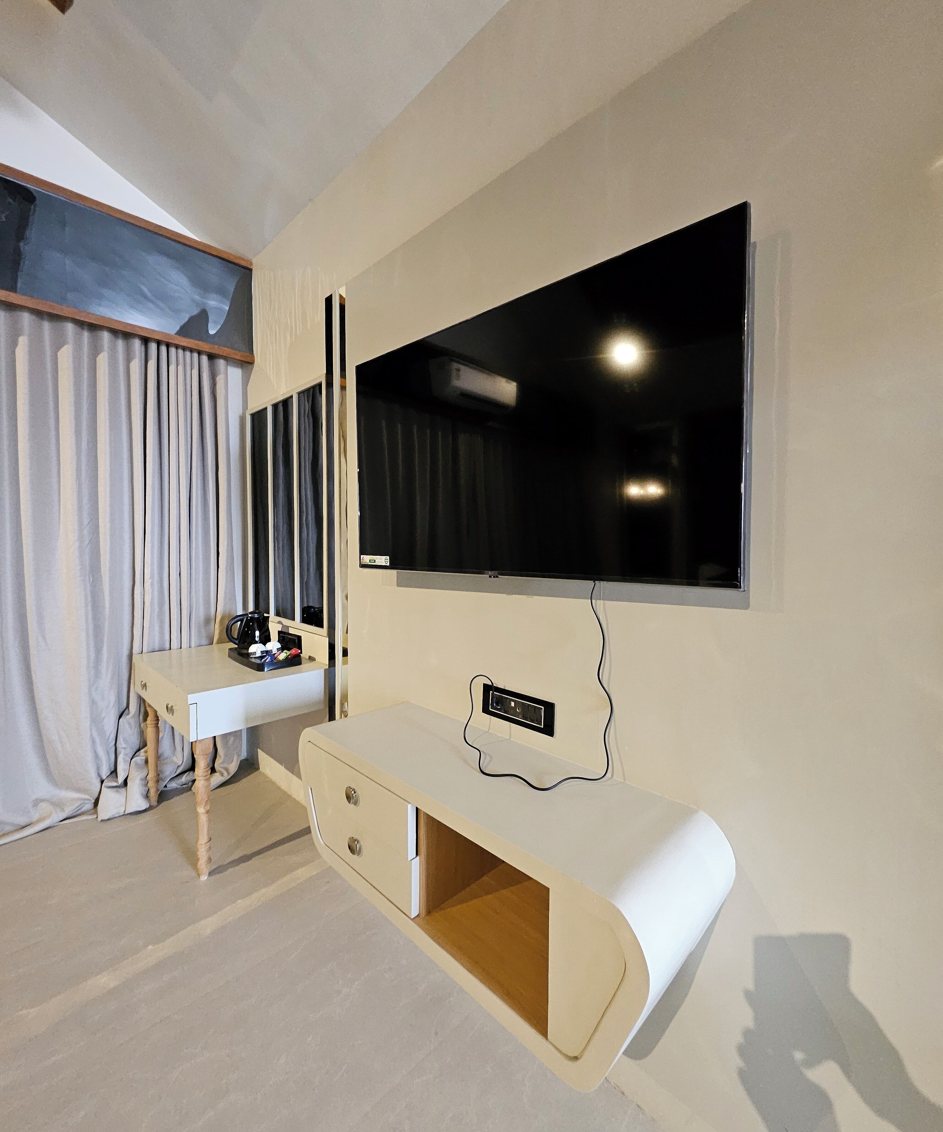 Een moderne hotelkamer met een grote flatscreen-televisie aan de muur, een houten bureau met lades, een klein krukje en een raam met gordijnen.