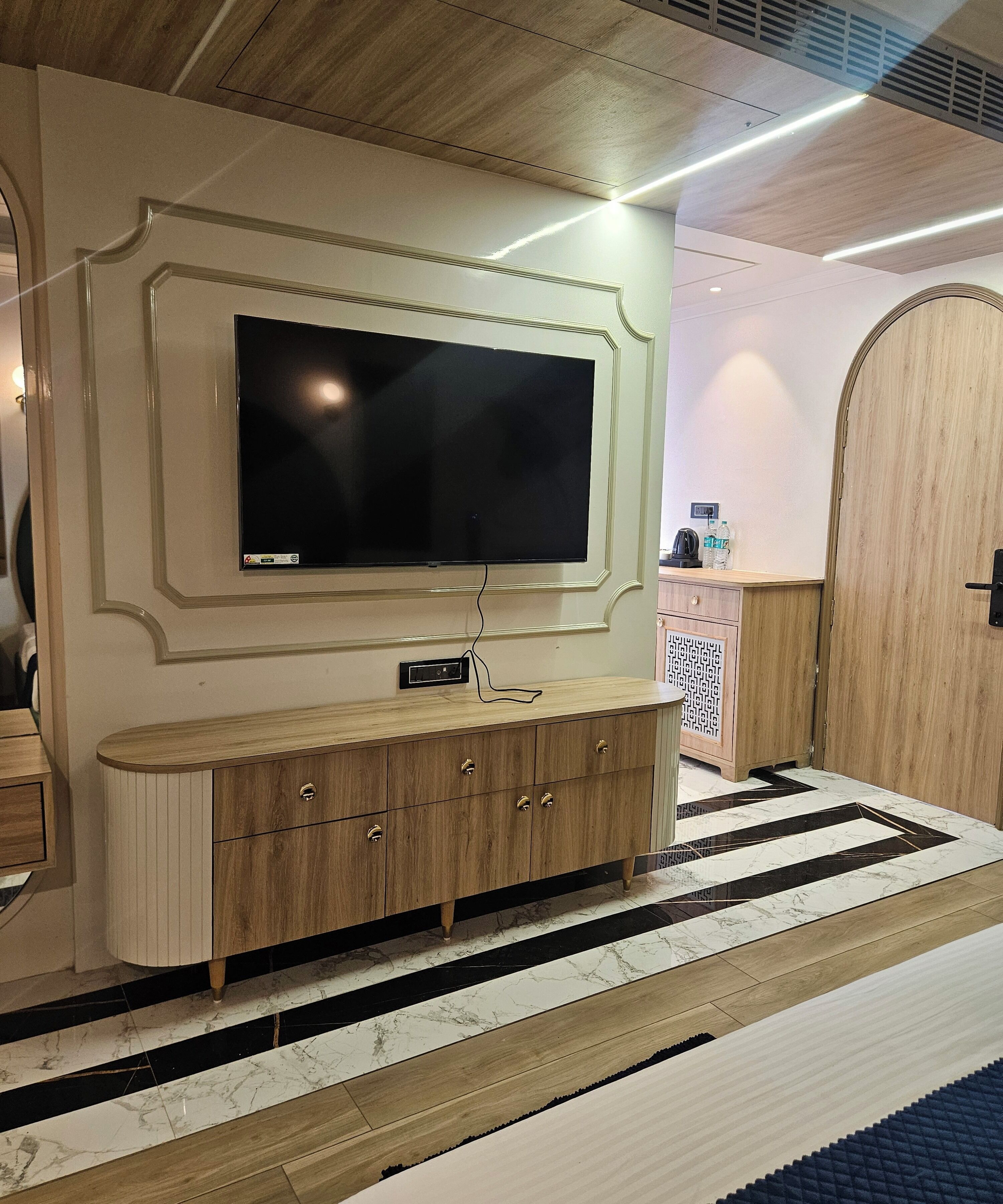 A modern hotel room with a large flat-screen TV mounted on a decorative wall, a wooden console table with drawers, and a patterned rug on the floor.
