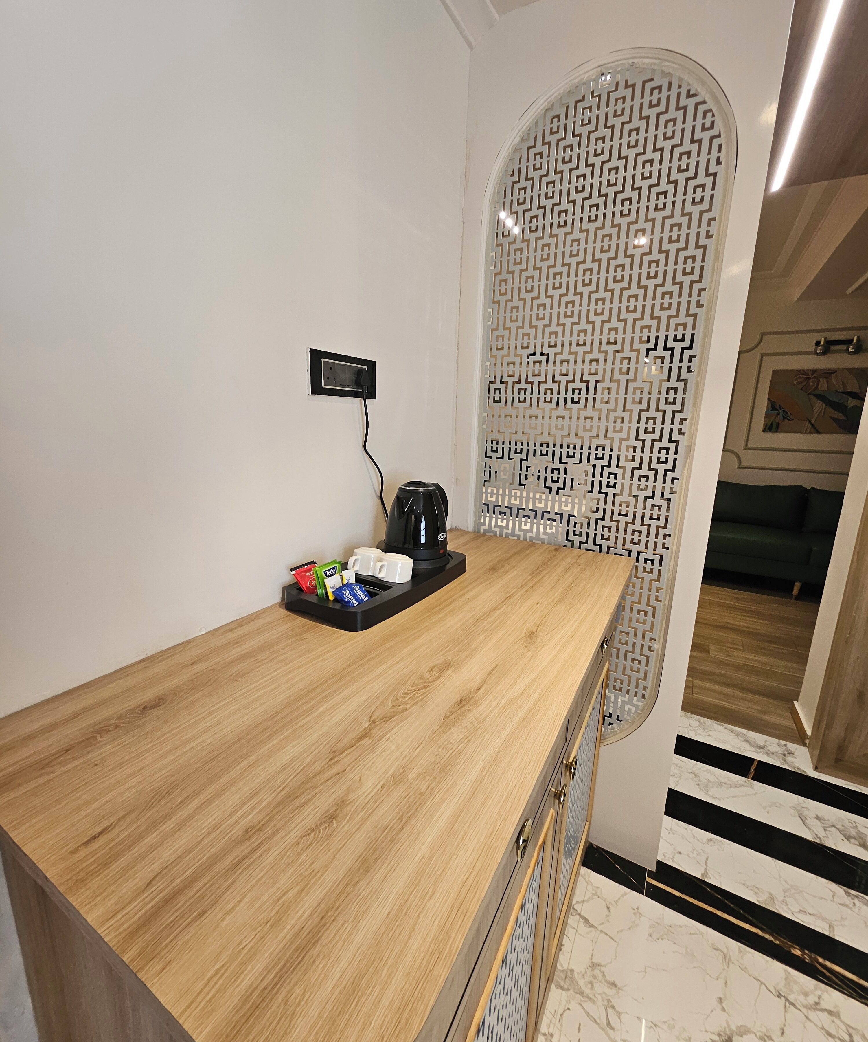 A wooden counter with a coffee maker and cups, a patterned wall, and a glimpse of a green couch and a hallway with a black and white striped floor.
