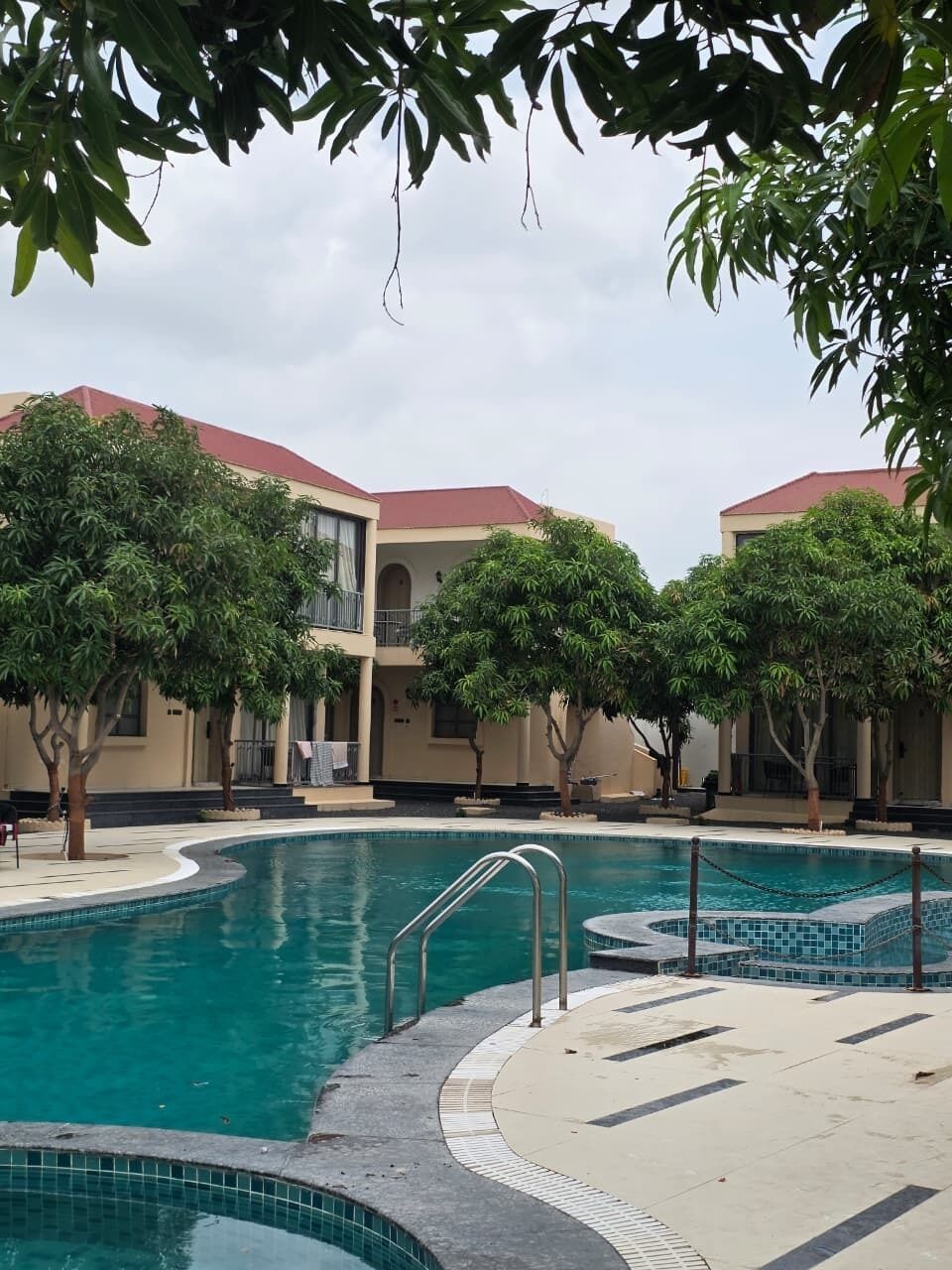 A swimming pool with a tiled deck, surrounded by trees and a building with a red roof.