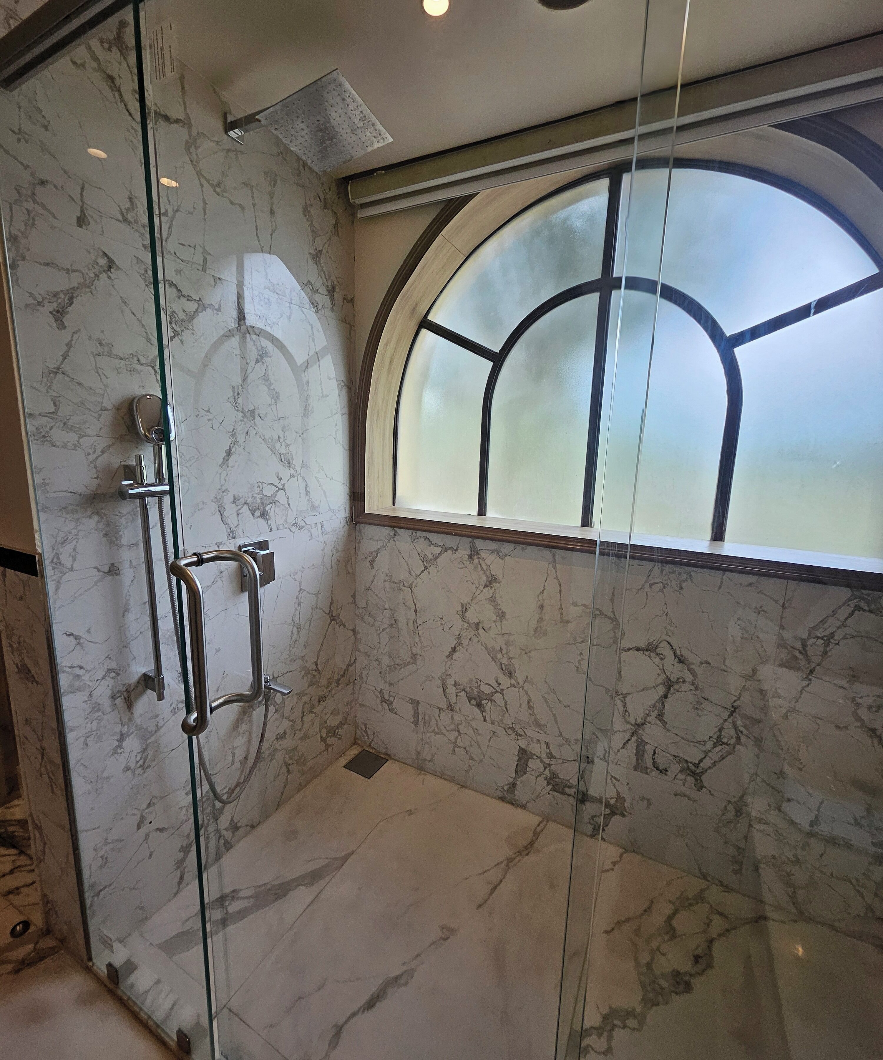 A glass-enclosed shower with a marble wall and a window with a curved design.