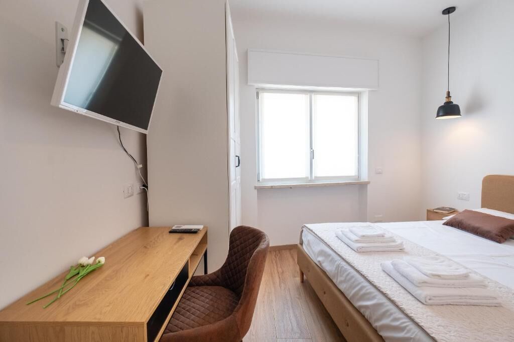 A modern hotel room with a bed, a desk, a chair, a TV mounted on the wall, and a window providing natural light.