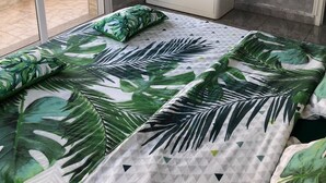 Une chambre à coucher avec un lit orné d’un motif de feuilles tropicales, une armoire blanche et une porte-fenêtre coulissante donnant sur un espace extérieur.
