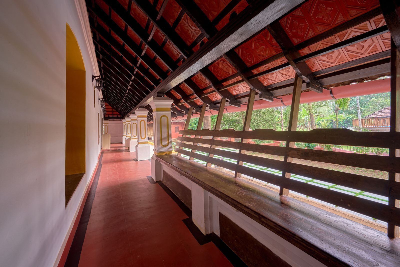 A corridor with a red-tiled floor, white pillars, and a wooden bench.