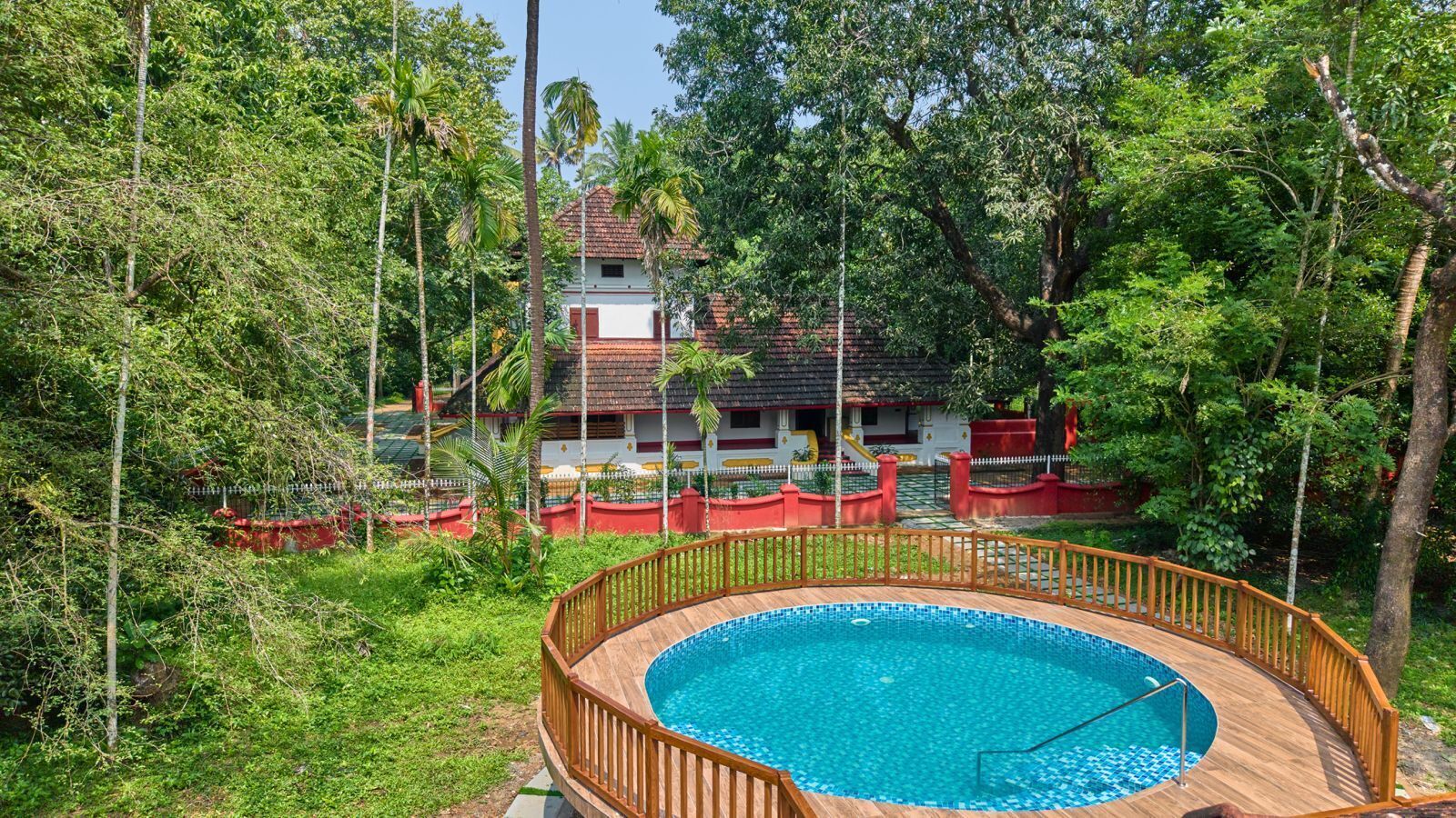 A swimming pool with a wooden deck, surrounded by lush greenery and a house with a red roof in the background.