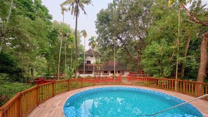 Ein runder Pool mit Holzdeck und Holzzaun, umgeben von üppiger Vegetation und einem Gebäude im Hintergrund.