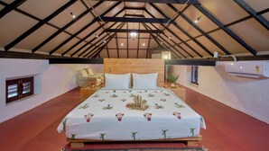 Ein Schlafzimmer mit hohen Decken, Holzbalken, einem Bett mit floralem Überwurf, einem Nachttisch aus Holz, einem Korbsessel und einem kleinen Fenster.