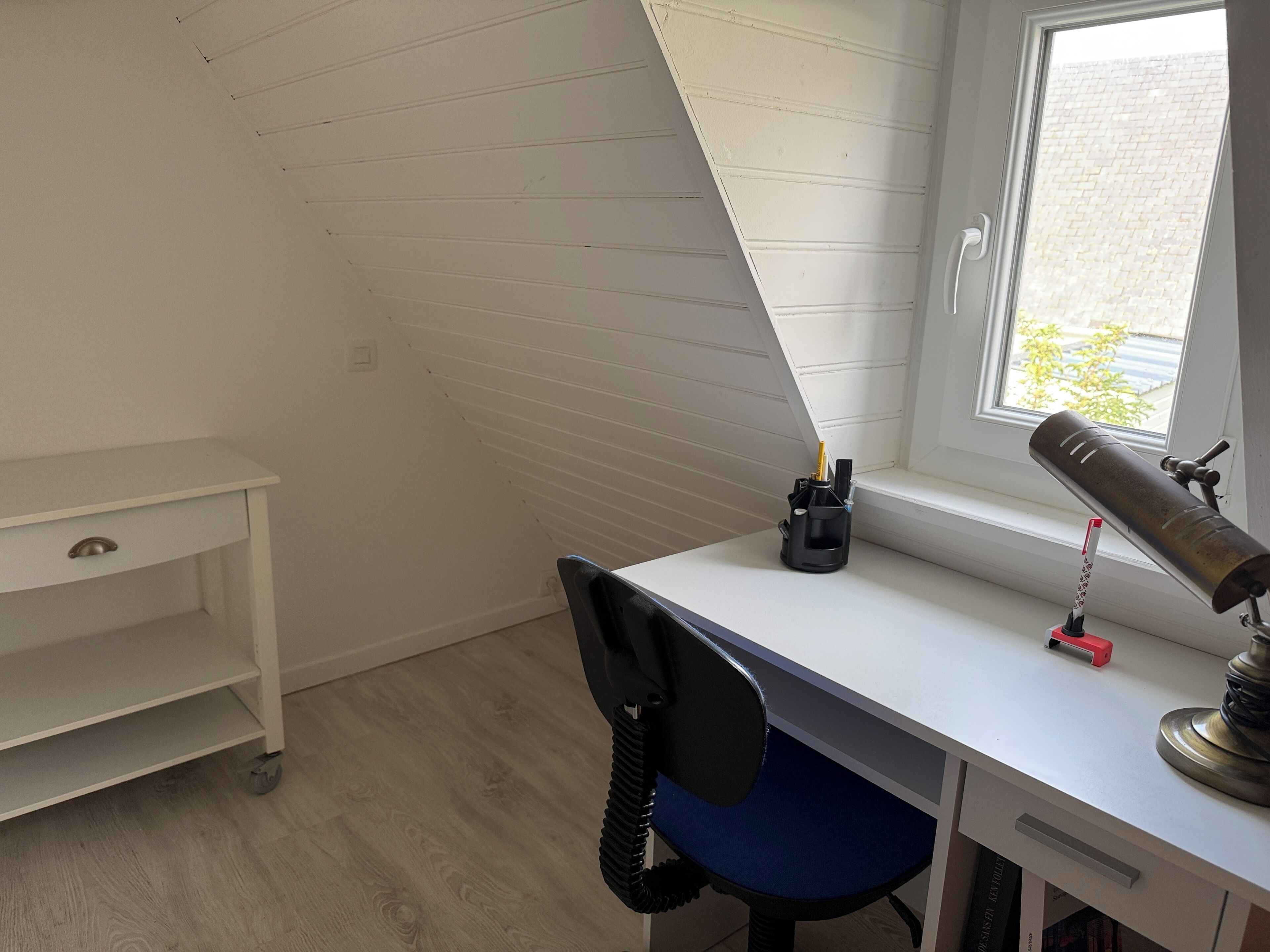 Ein Zimmer mit Schreibtisch, Stuhl, Lampe und einem Fenster mit Ausblick nach draußen.