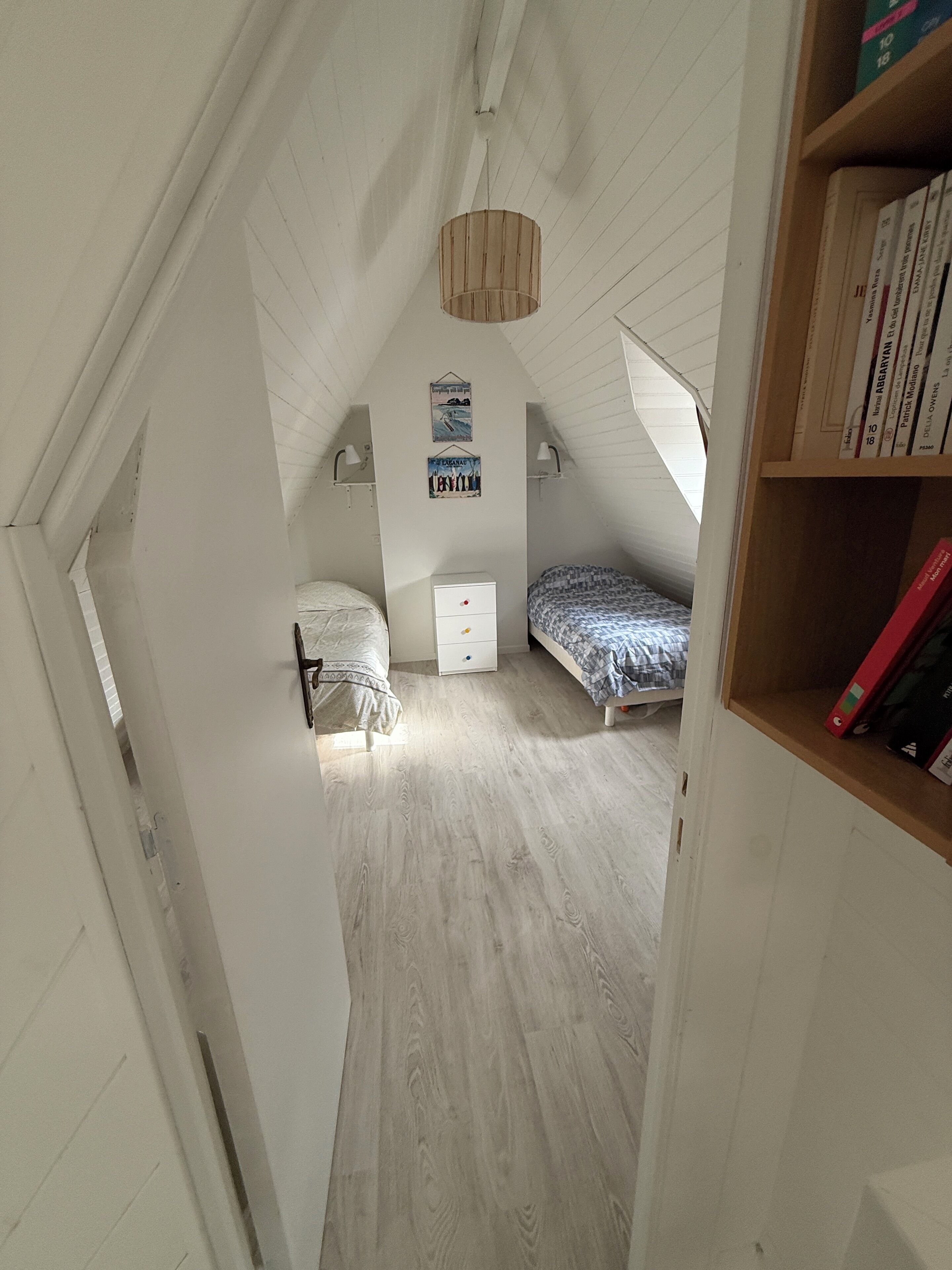 Un pequeño dormitorio en el ático con techos inclinados, dos camas, un tocador blanco y una estantería de madera.