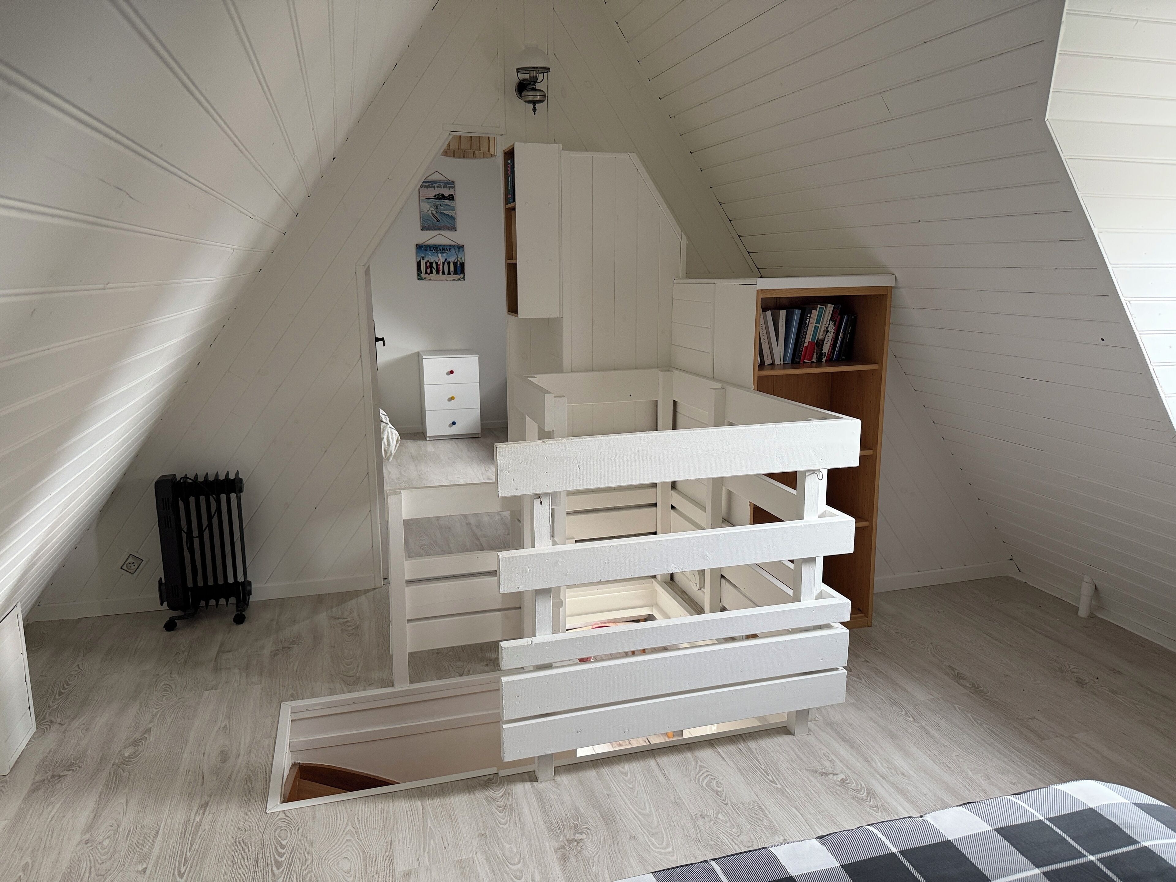 Une pièce avec un plafond en pente, un lit blanc avec des tiroirs, un radiateur, une bibliothèque et une petite fenêtre.