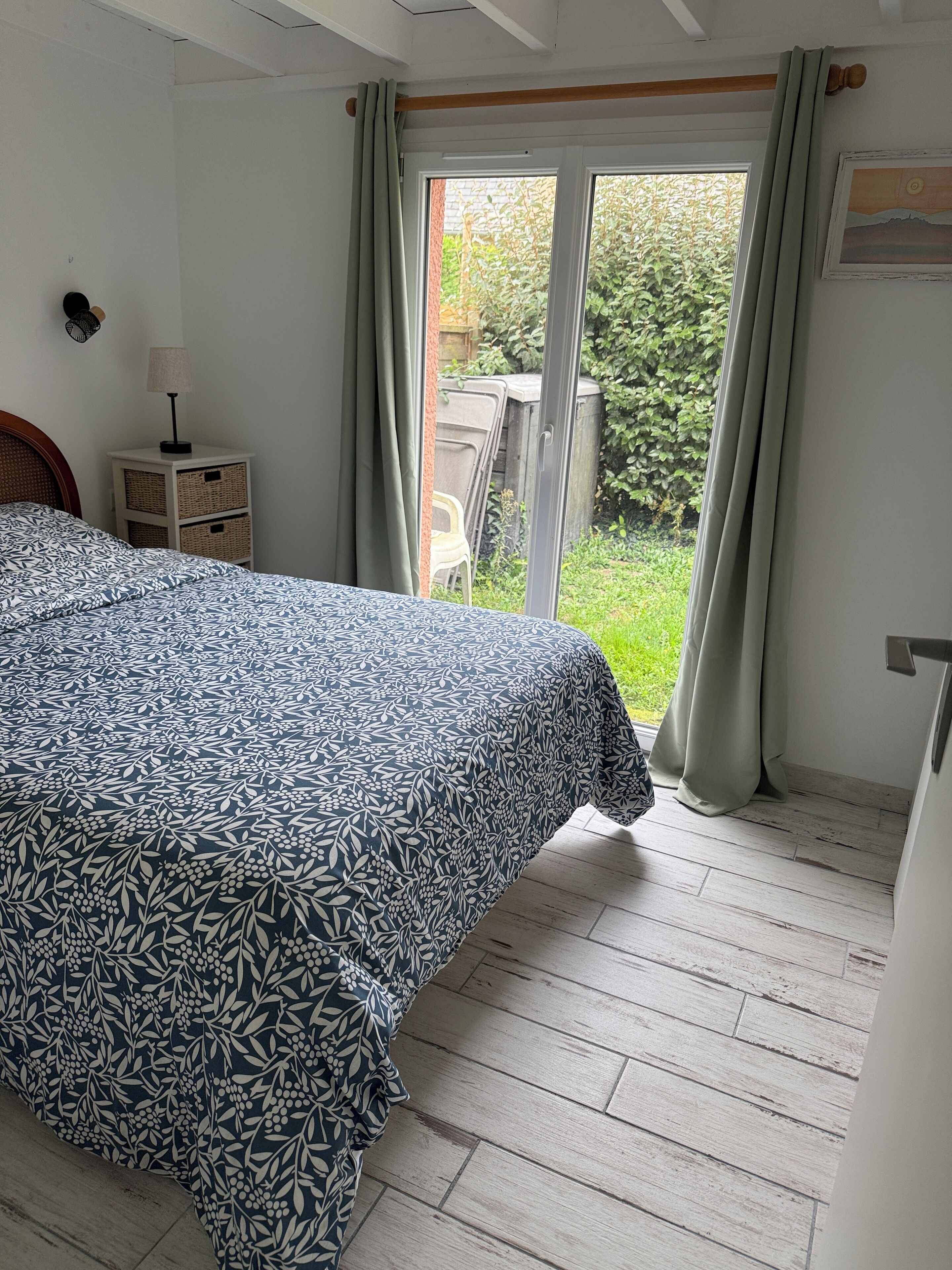 A bedroom with a bed, a nightstand, a window with curtains, and a view of greenery outside.