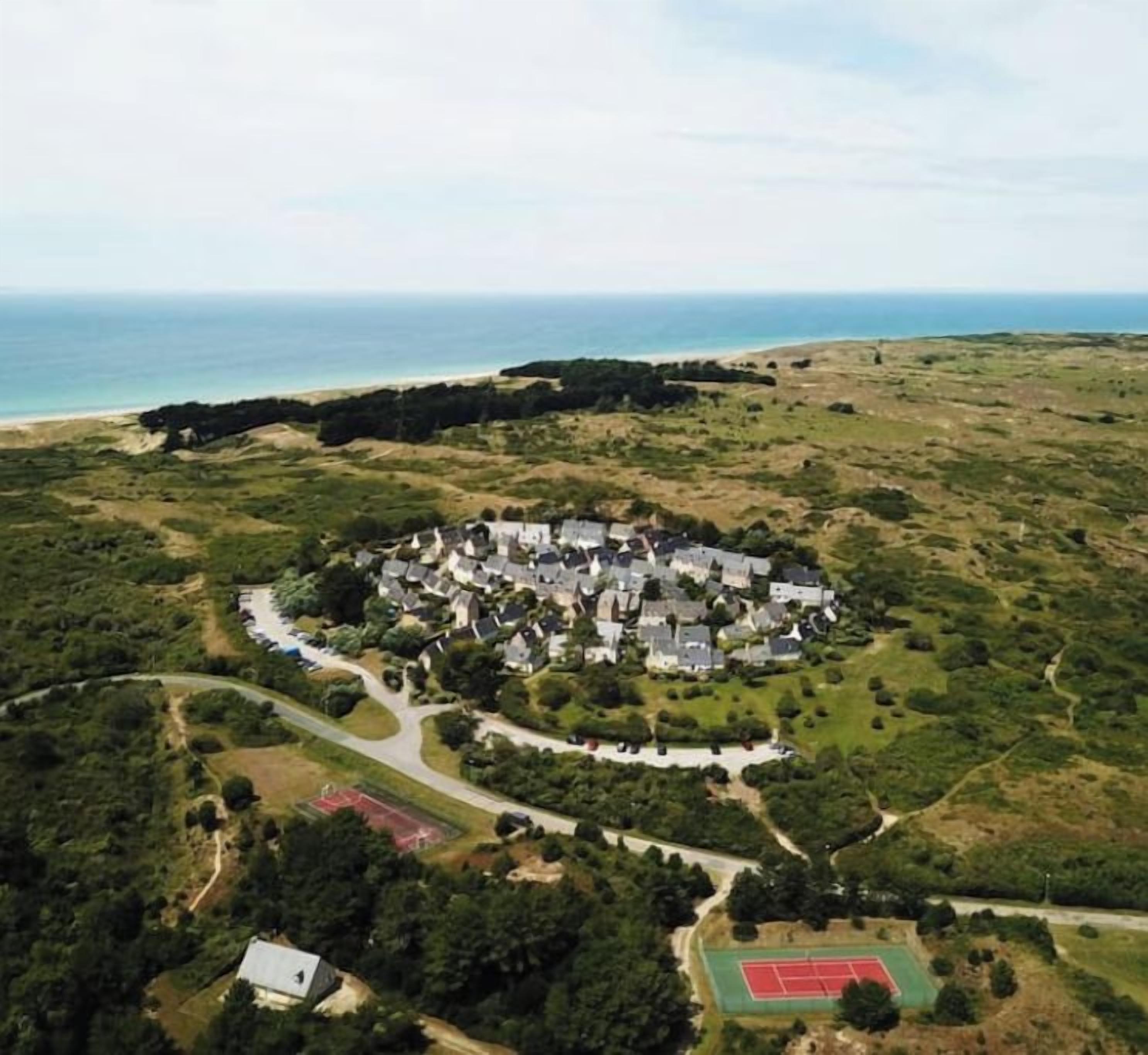 Vue aérienne d’un quartier résidentiel avec des maisons, un court de tennis et une plage en arrière-plan.