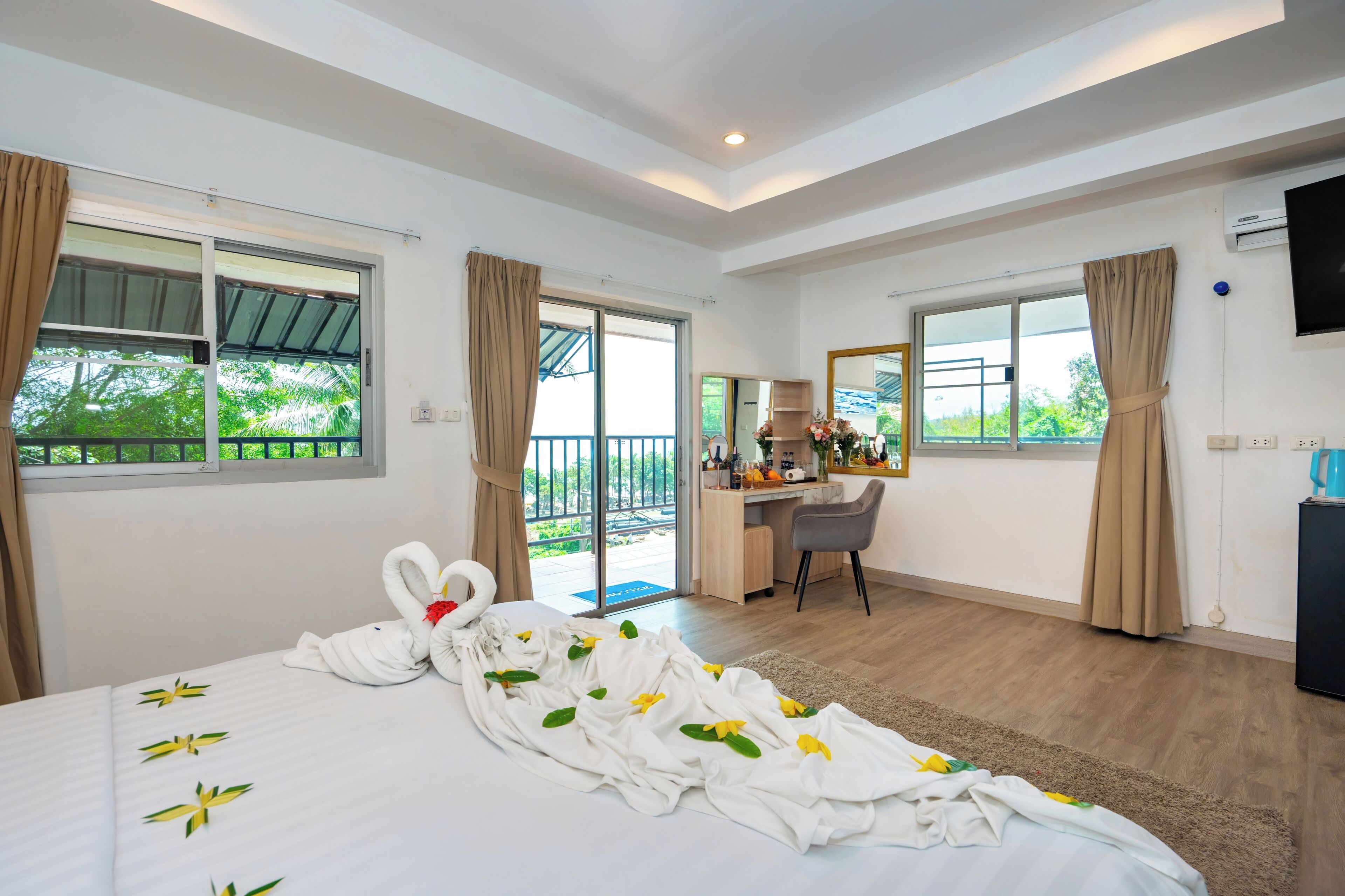 A hotel room with a bed, a desk, a chair, a TV, and a balcony with a view of greenery.