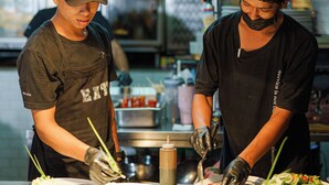 Dos cocineros preparando comida en una cocina con iluminación industrial y electrodomésticos de acero inoxidable.