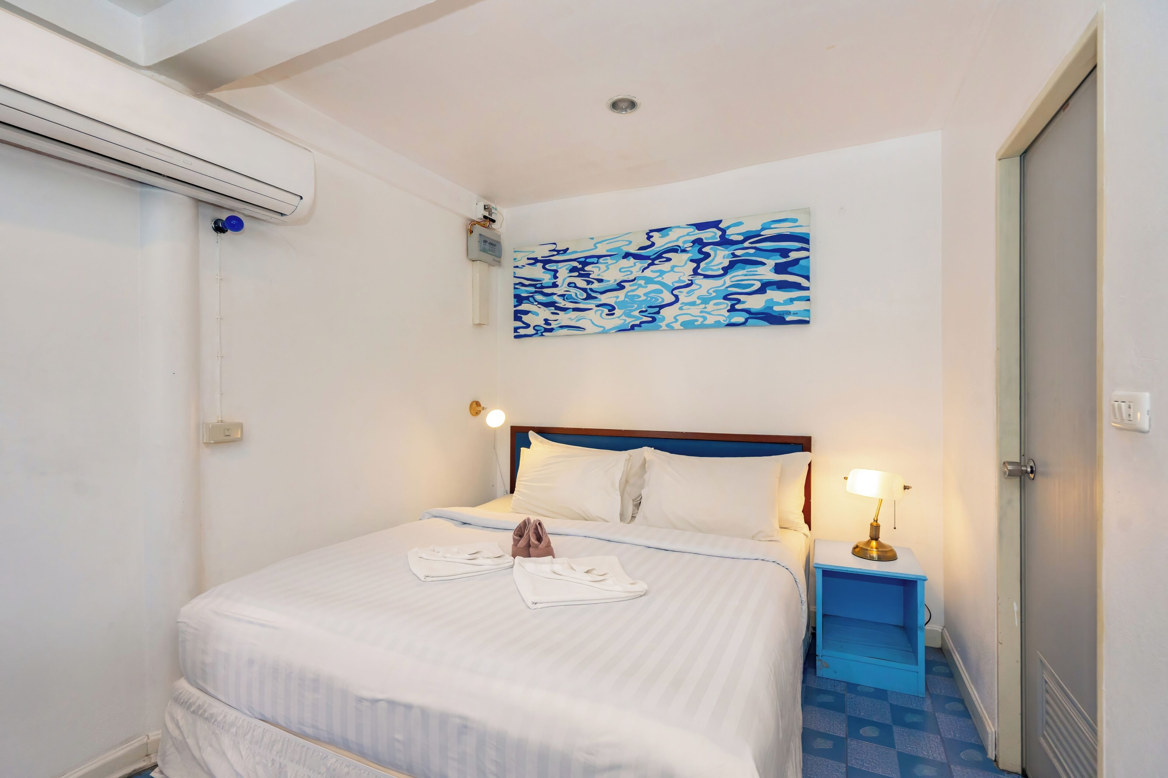 A hotel room with a bed, a painting, a bedside lamp, and a blue bedside table.