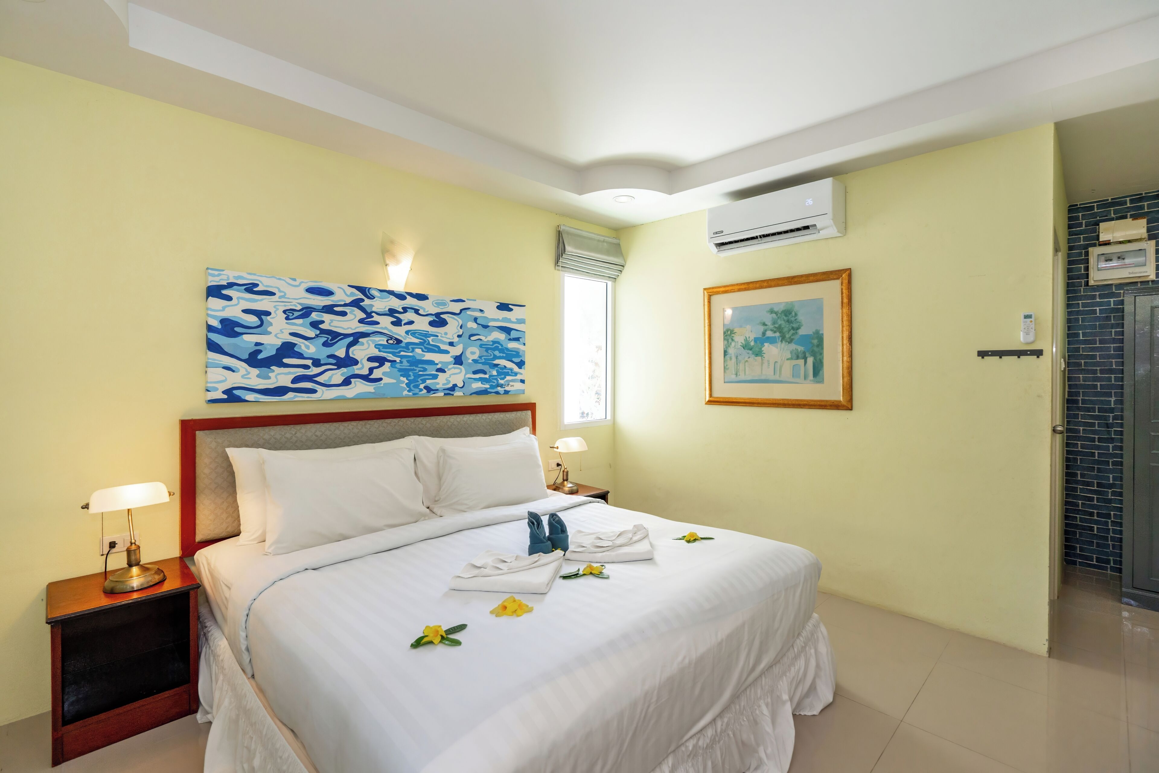 A hotel room with a bed, bedside tables, a wall-mounted air conditioner, a framed picture, and a window with a view.