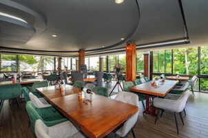 A modern restaurant with wooden tables and green upholstered chairs, large windows offering a view of greenery, and a curved ceiling design.