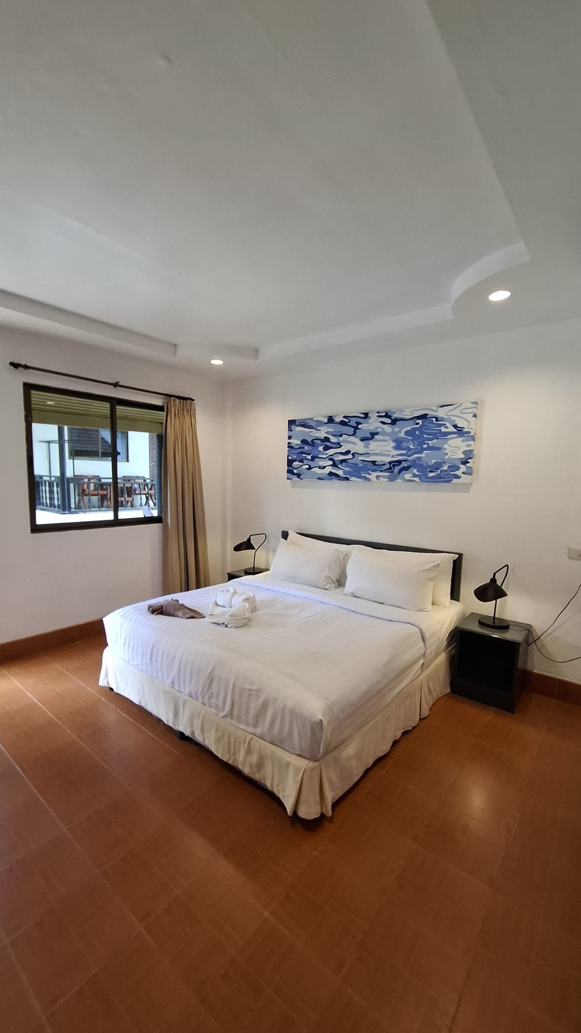 A hotel room with a large bed, a nightstand, a lamp, a window with curtains, and a painting on the wall.