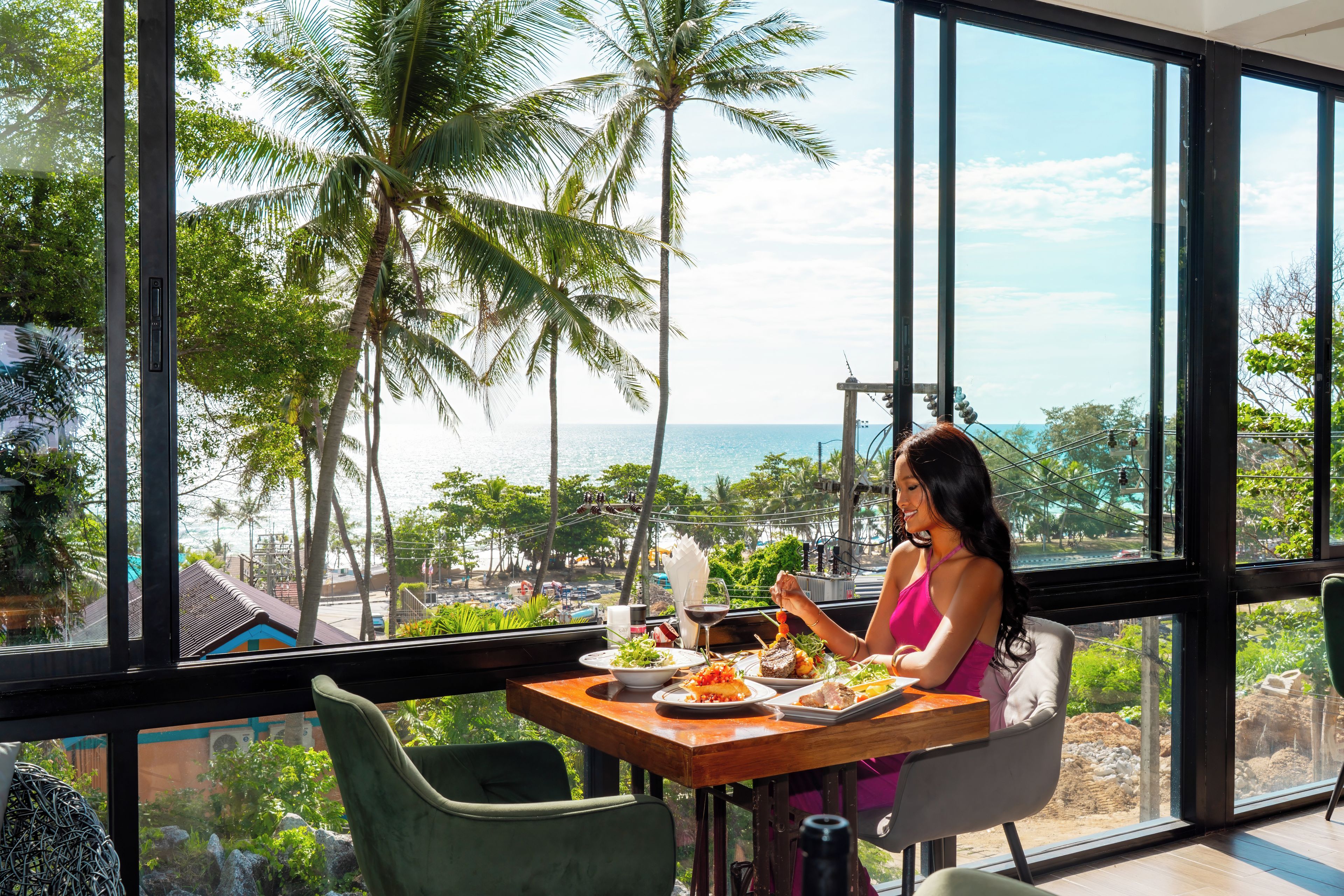 A dining area with a view of the sea and palm trees, featuring a table with plates of food and a person dining.