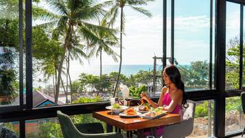 A dining area with a view of the sea and palm trees, featuring a table with plates of food and a person dining.
