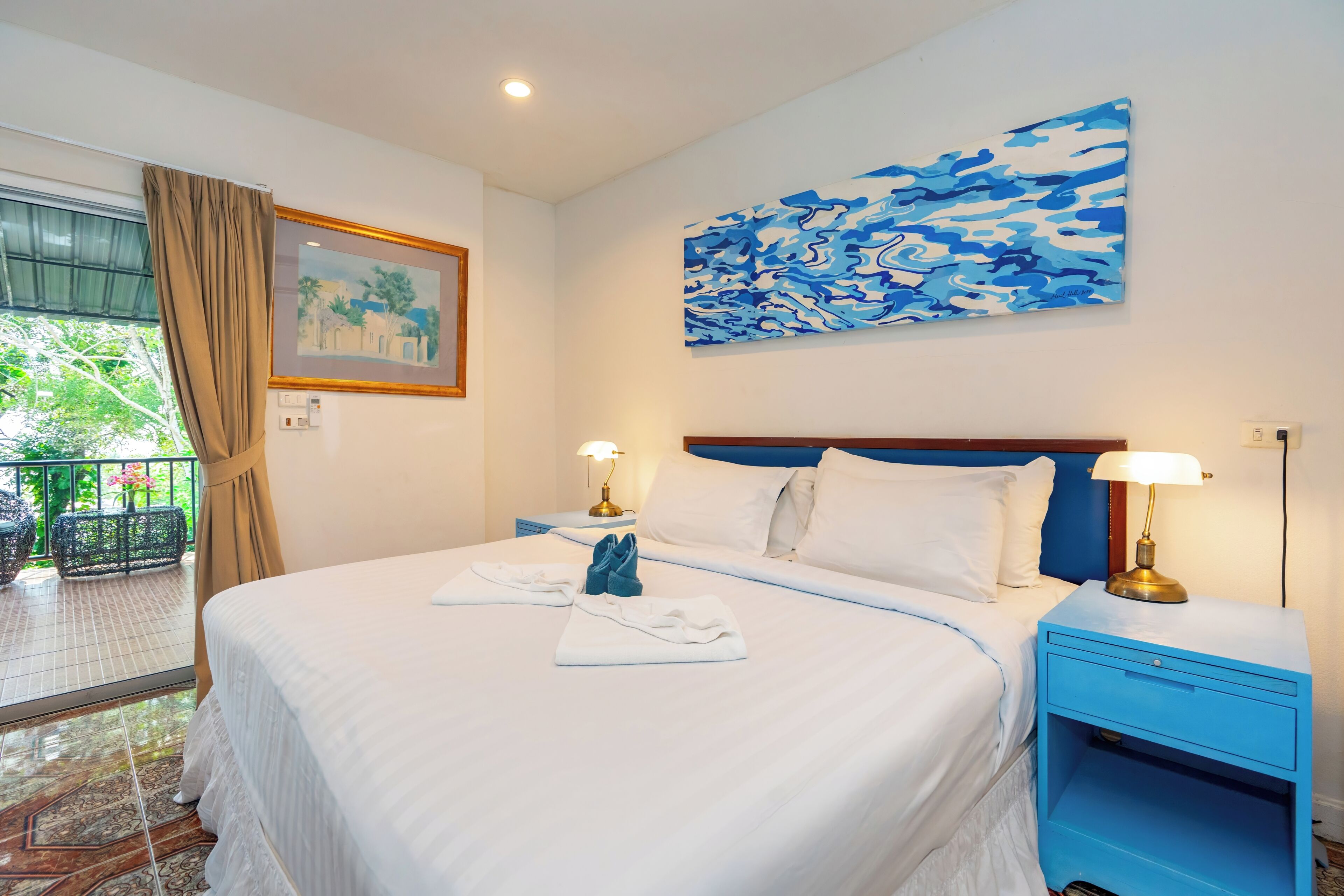 A hotel room with a bed, two bedside tables, a painting, and a view of greenery outside.