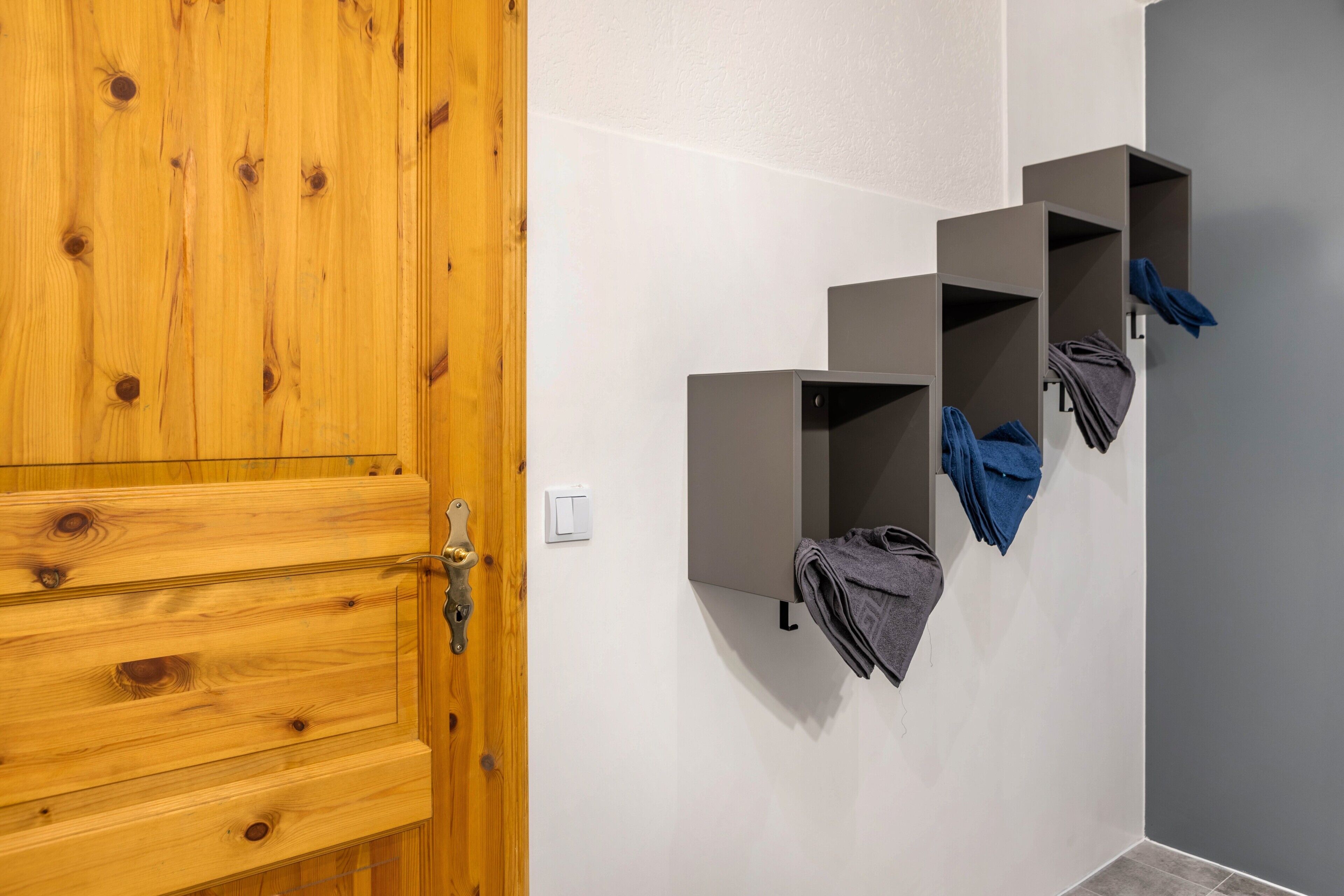 A wooden door with a brass handle, a white wall with a light switch, and three grey storage units with folded towels.