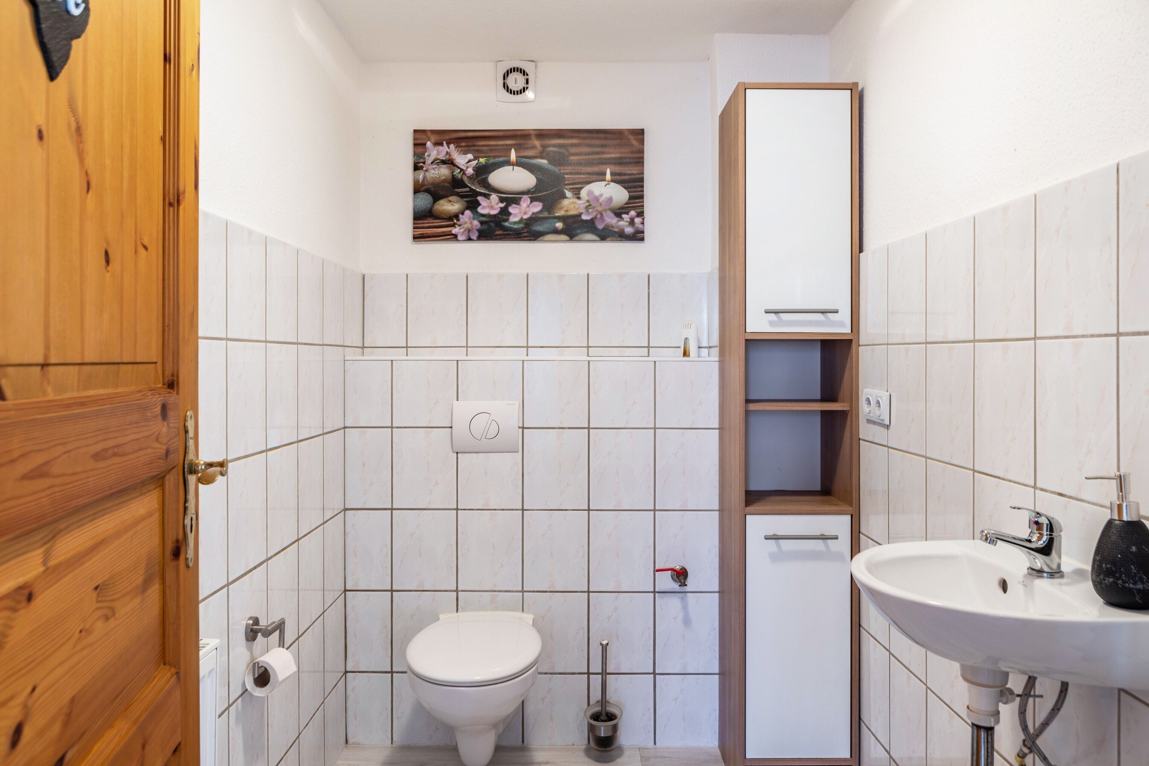 Un bagno rivestito di piastrelle bianche, con il water, un lavandino, una porta in legno e uno scaffale a parete.