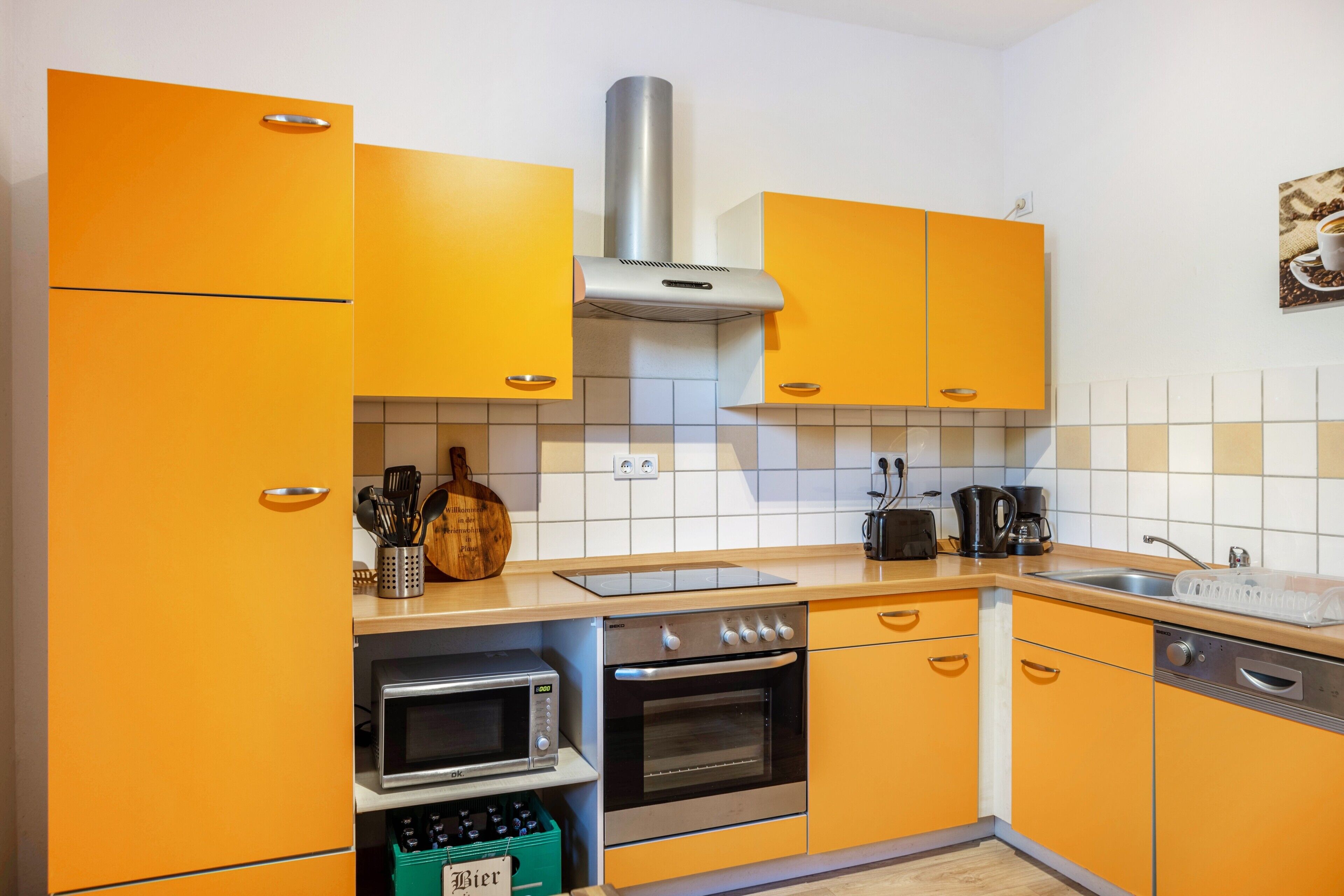 Une cuisine avec des meubles de couleur orange, une hotte de cuisinière en acier inoxydable, un four à micro-ondes, un four traditionnel et un évier.