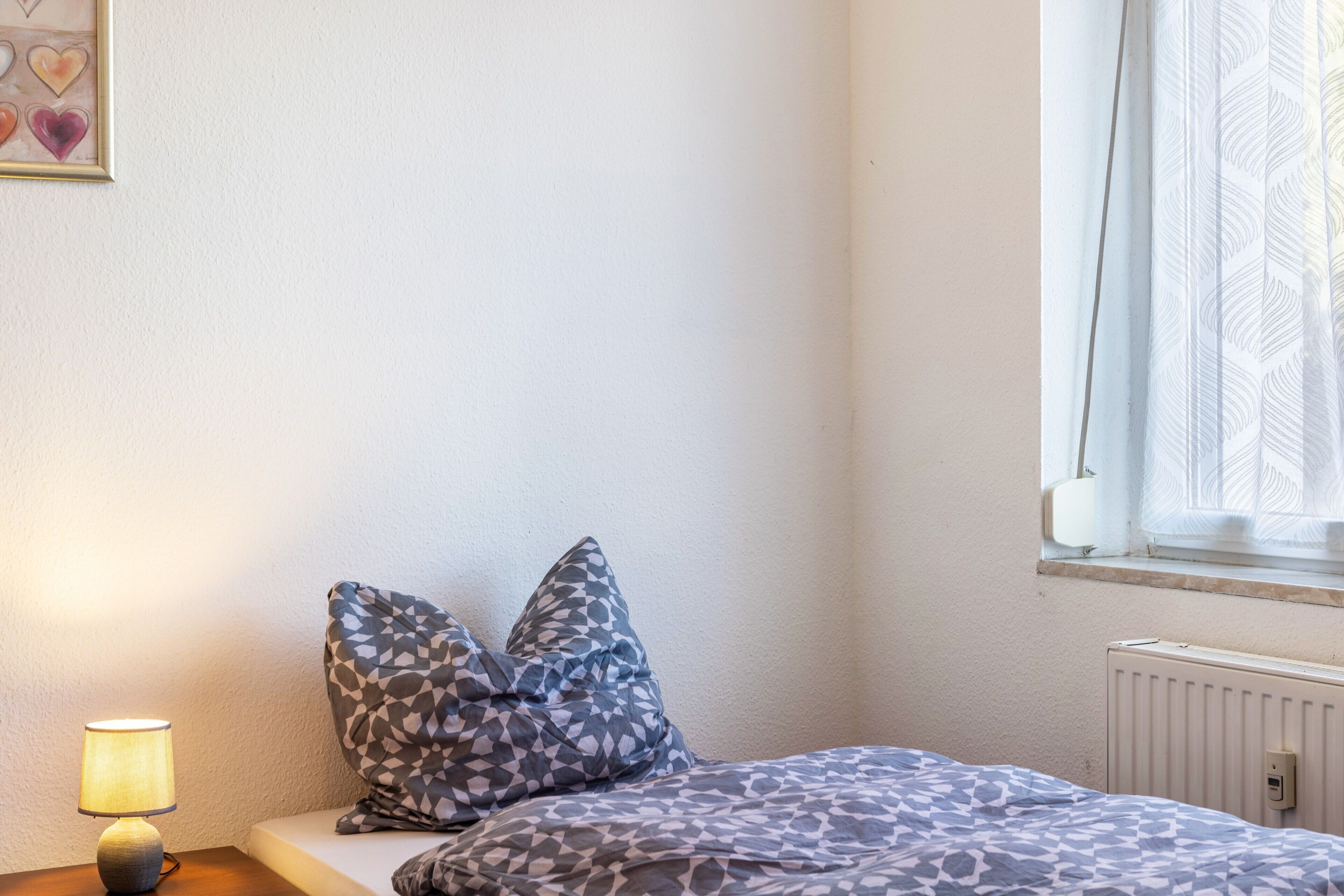 Une chambre à coucher avec un lit, des oreillers, une lampe, un radiateur et une fenêtre avec des rideaux.