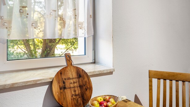 Uma janela com cortina branca, tábua de corte de madeira com texto e uma cesta de frutas sobre uma mesa.