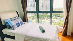 A neatly made bed with a white sheet and two pillows, a rolled-up blanket, and a view of tall buildings and greenery through large windows. - Teega Residence (Iskandar Puteri)
