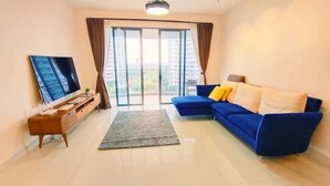 A modern living room with a flat-screen TV, a blue sofa with cushions, a small wooden side table, and a large sliding glass door leading to an outdoor balcony. - Teega Residence (Iskandar Puteri)