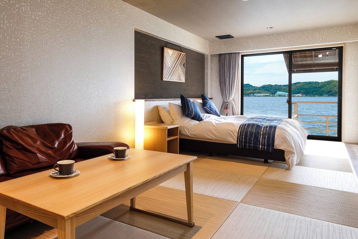 A hotel room with a wooden table, a leather sofa, a bed with a view of the water, and a large window.