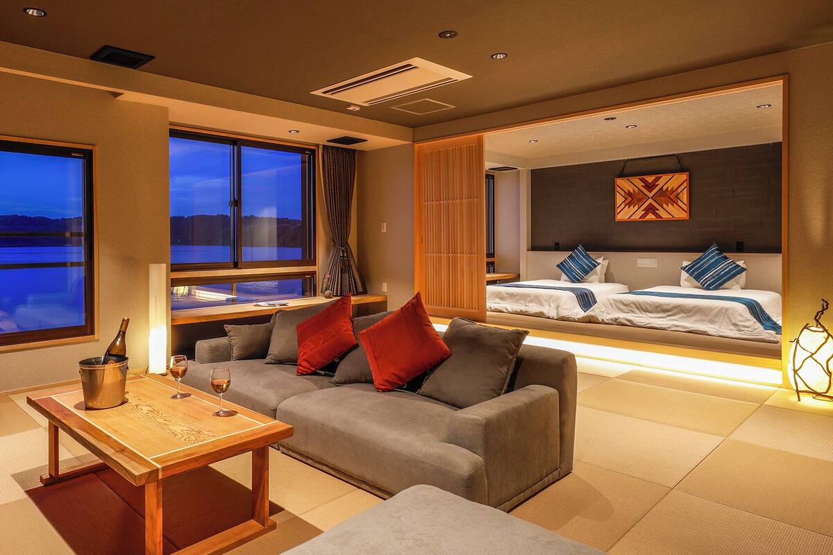 A modern hotel room with a large window offering a scenic view, a cozy seating area with a wooden table, and a separate bedroom with a bed and a decorative wall piece.
