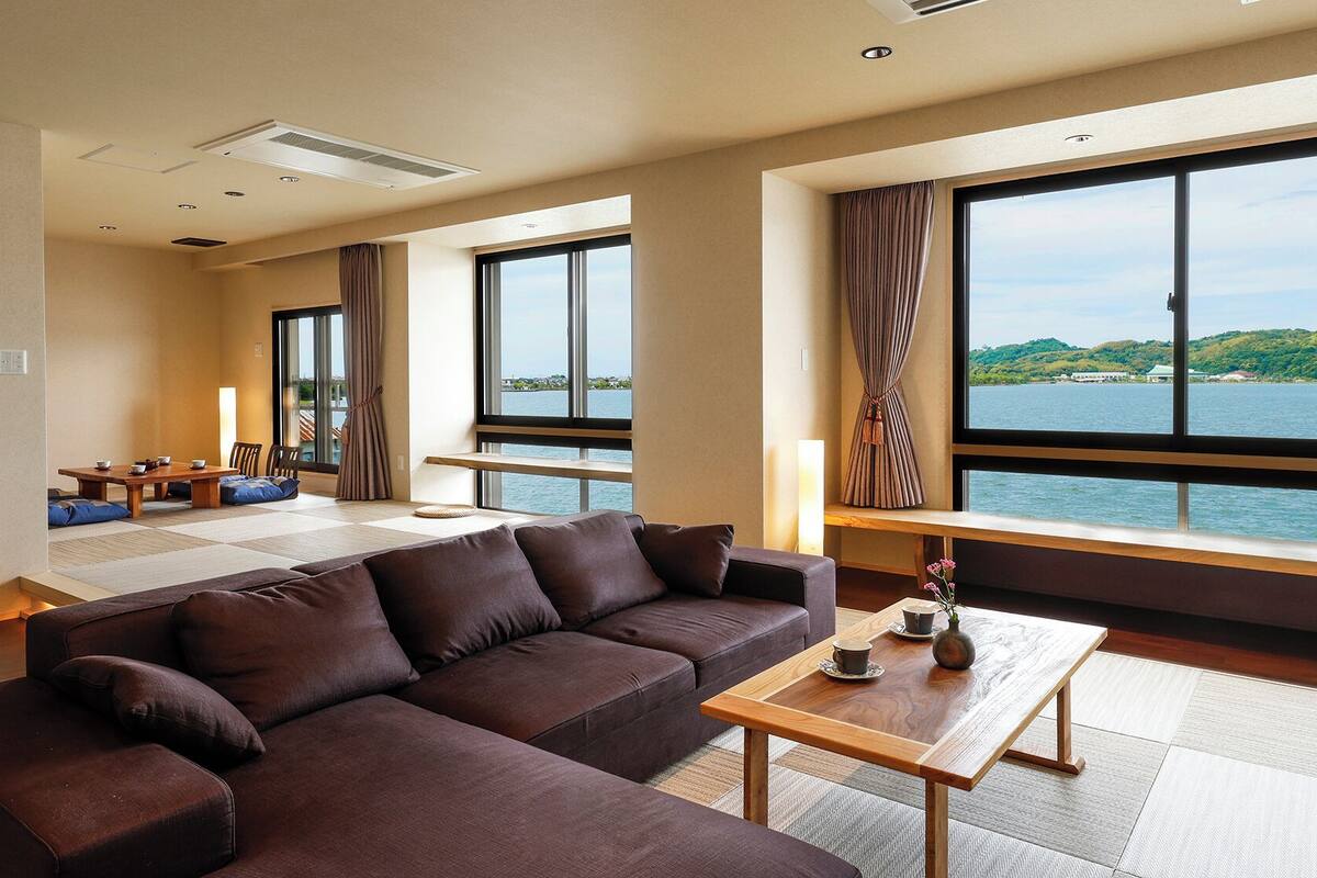 A modern hotel room with a large sofa, a dining area, and a view of a lake.