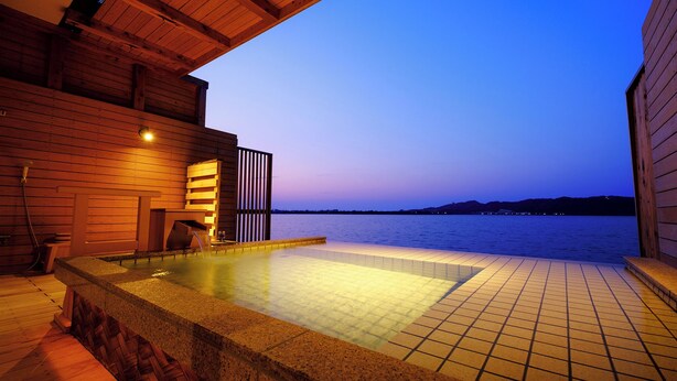 A wooden spa area with a hot tub and a view of the water at dusk. - Sennentei (Yurihama)