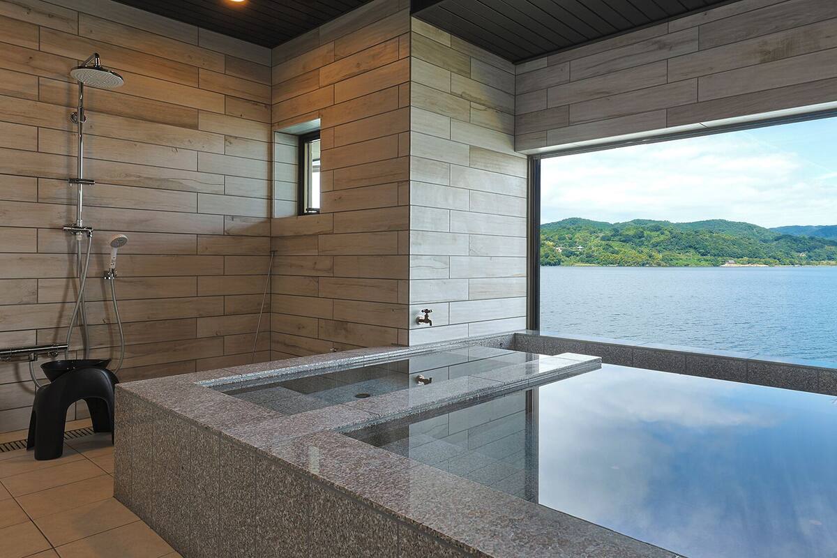 A modern bathroom with a glass-enclosed hot tub, wooden walls, and a view of a lake.