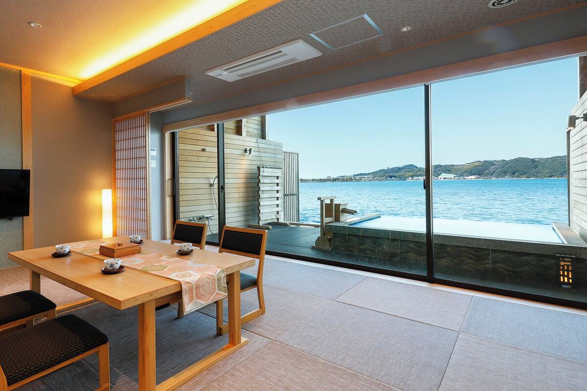 A modern room with a wooden table and chairs, a flat-screen TV, and a large window overlooking a body of water.