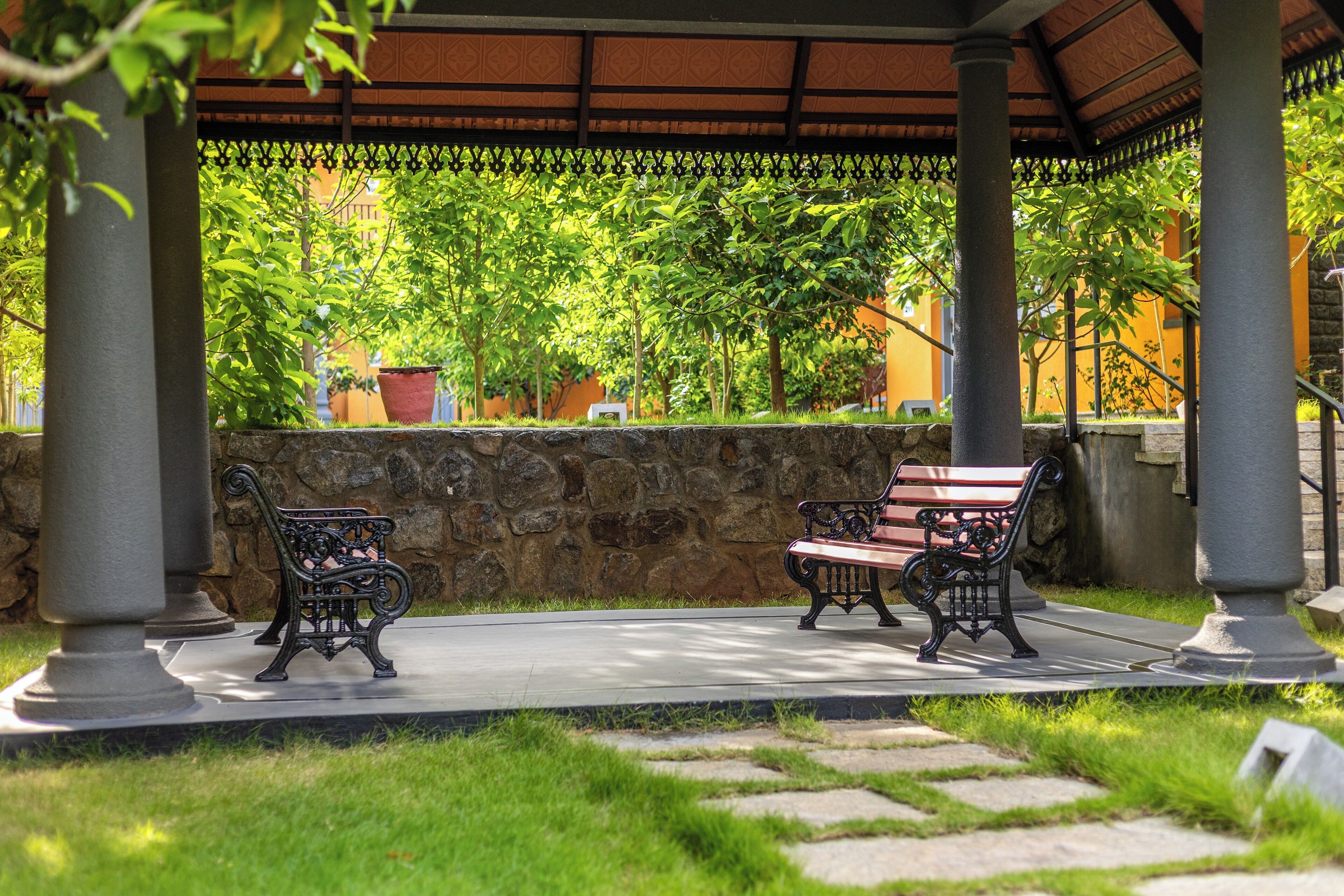 A covered outdoor seating area with two ornate benches, a stone wall, and lush greenery.