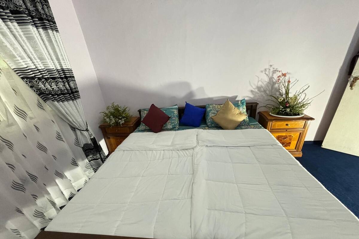 A bed with a white mattress, a wooden headboard, and a blue carpet. There are several pillows in various colors and patterns, and a wooden nightstand with a plant on it.