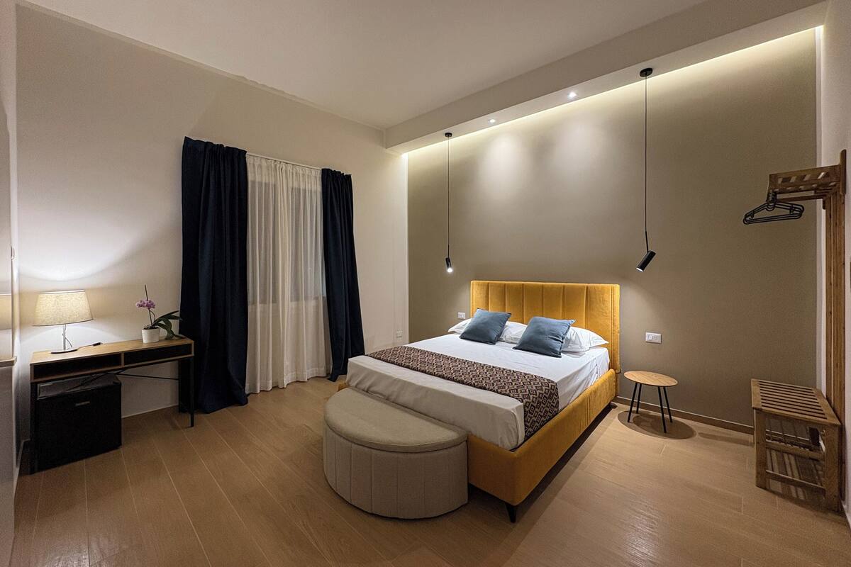 Moderni hotellihuone, jossa on sänky, yöpöytä, lamppu ja pieni pyöreä pöytä.