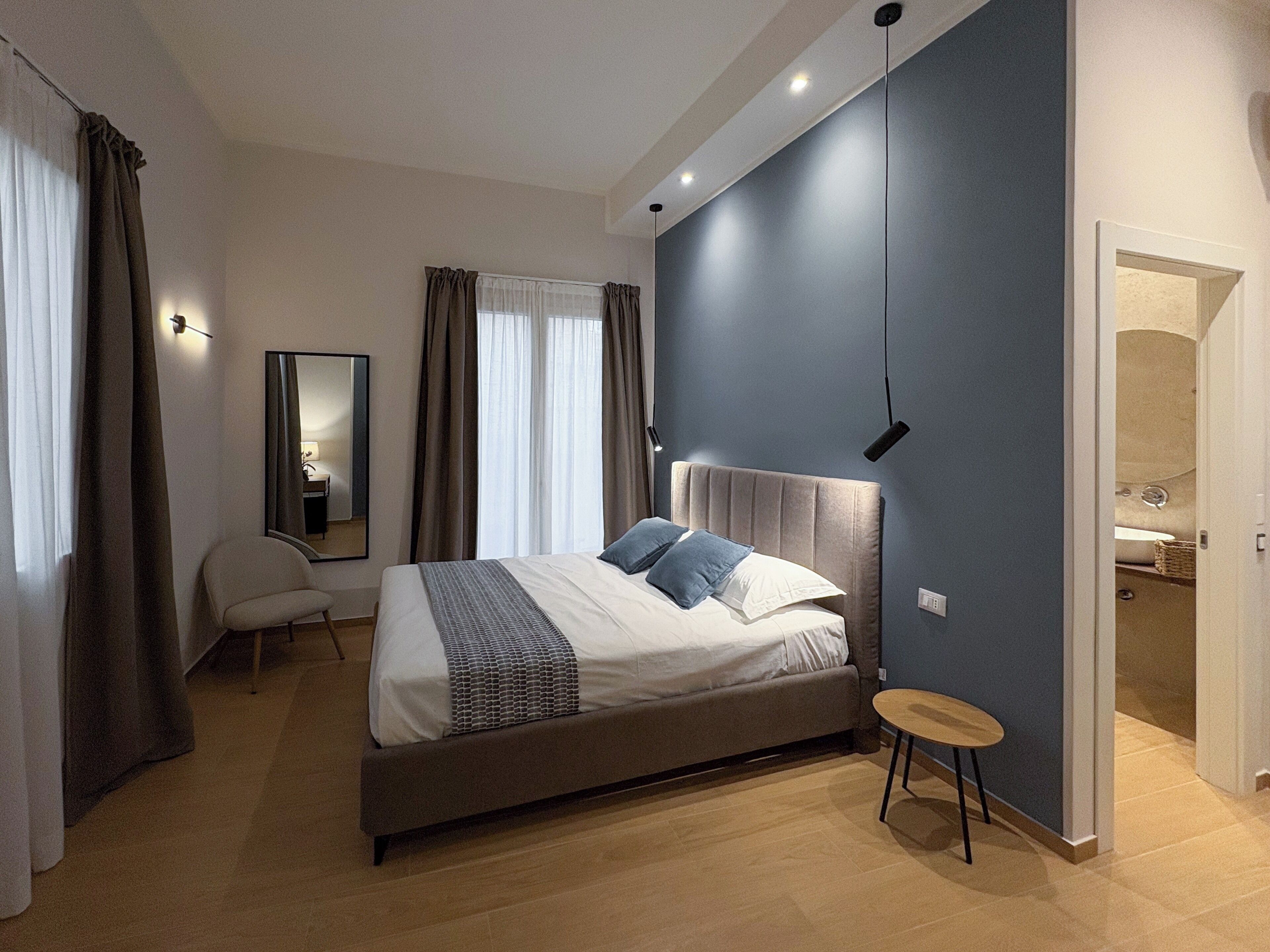A modern bedroom with a large bed, a bedside table, a chair, a mirror, and a bathroom visible through an open door.