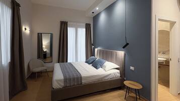 A modern bedroom with a large bed, a bedside table, a chair, a mirror, and a bathroom visible through an open door.