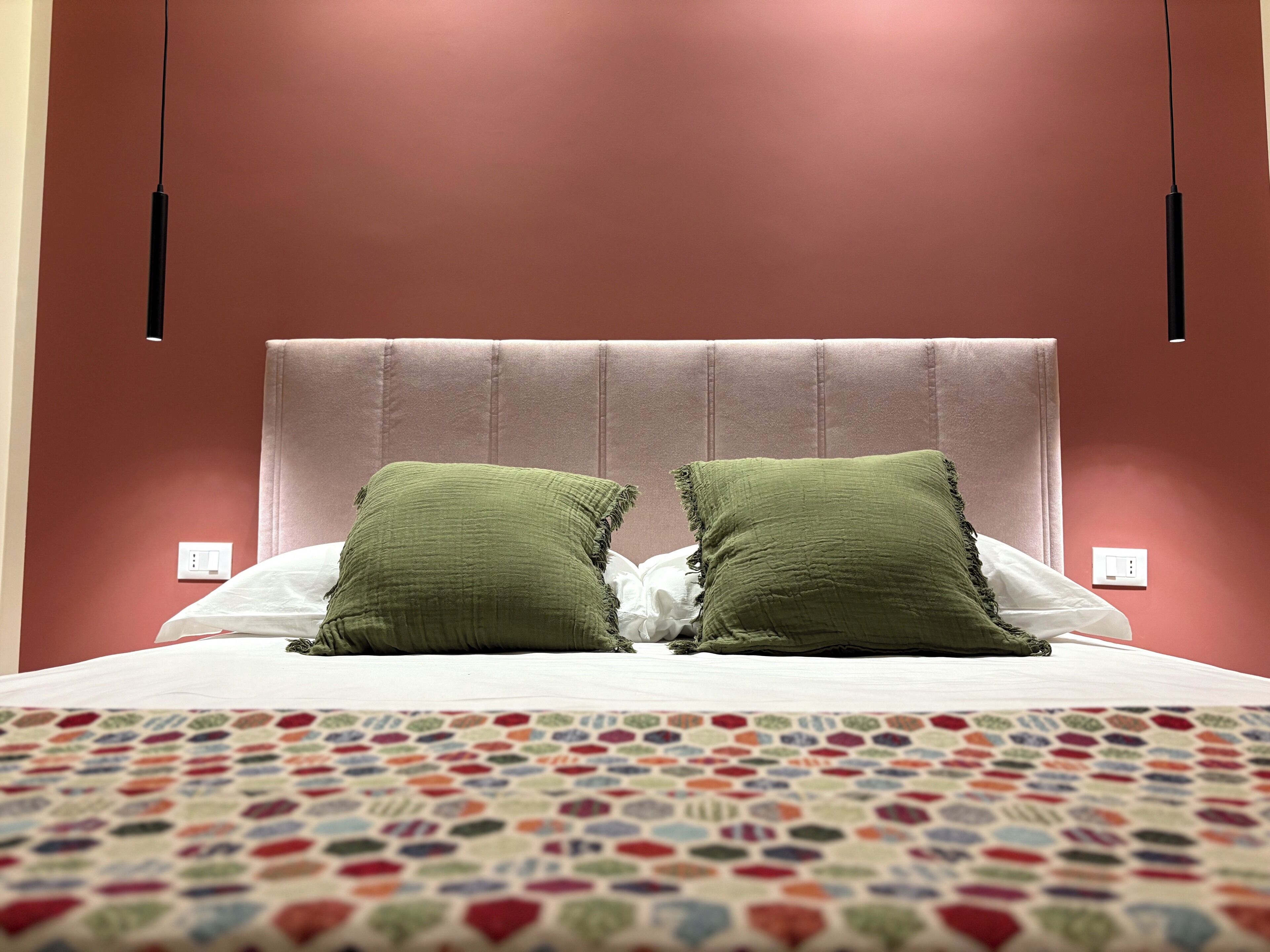 A bed with a patterned bedspread, two green pillows, and a headboard against a red wall.